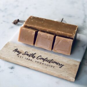 1 bar of clotted cream fudge with 3 slices of clotted cream fudge leant against it. It is sat on a marble & wood board with the text 'Amy Smiths Confectionery, est 1986, Yorkshire'.